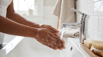 Primer plano de manos femeninas de piel morena enjabonándose bajo el agua de un grifo cromado en un lavabo blanco de un baño aseado.