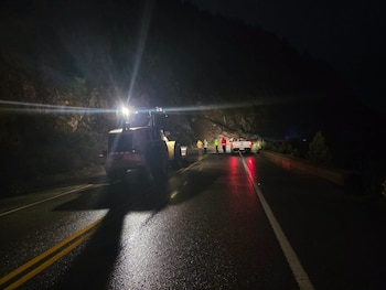 Una cargadora frontal amarilla, un vehículo pickup blanco, varias personas con chalecos reflectantes, una carretera mojada y una ladera rocosa de noche
