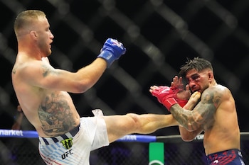 Dos luchadores en la jaula de UFC. Uno, con shorts blancos, patea al otro, con guantes rojos y el rostro ensangrentado. Se ven tatuajes y la malla de la jaula