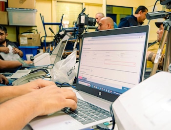 Manos tecleando en un computador portátil HP con un formulario en pantalla, un trípode con cámara, varias personas, cajas y una grapadora