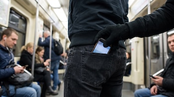 Una mano con guante negro saca un teléfono celular del bolsillo trasero del pantalón de un hombre dentro de un metro con pasajeros.