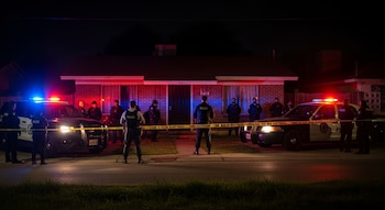 Dos coches de policía con luces rojas y azules intermitentes, cinta policial amarilla, varios agentes de policía y una casa de ladrillo por la noche.
