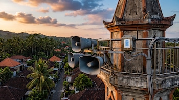 Tres altavoces metálicos y un sensor con la sigla del PNUD en la balconada de un minarete frente a un paisaje de casas y palmeras.