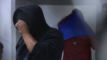 Un hombre con sudadera negra cubre su rostro con la mano, mientras otra persona detrás tapa su cabeza con una prenda azul
