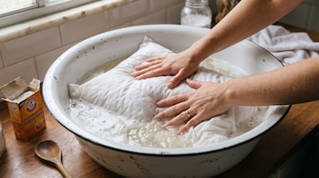 Manos lavando una almohada blanca en un recipiente metálico con agua y espuma. Caja de bicarbonato de sodio y cuchara de madera sobre mesa de madera.