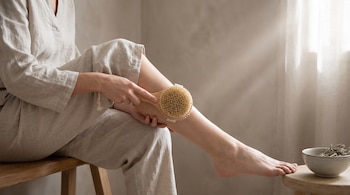 Mujer sentada en ropa ligera cepillando su pierna descubierta con un cepillo de cerdas naturales; un cuenco con hierbas secas se ve a su lado.