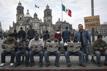 Seis hombres en un banco, algunos leyendo periódicos, otros con teléfonos. Un hombre de pie con un cartel: Necesito Trabajo. Al fondo, la Catedral Metropolitana y dos banderas de México.