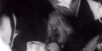 Imagen en blanco y negro borrosa de Hendrik Verwoerd con la boca abierta, rodeado por hombres que lo asisten tras un ataque, en un evento caótico