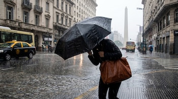 Persona con paraguas negro y bolso marrón caminando por una calle adoquinada mojada bajo la lluvia, con taxis, buses, edificios y el Obelisco de Buenos Aires al fondo.