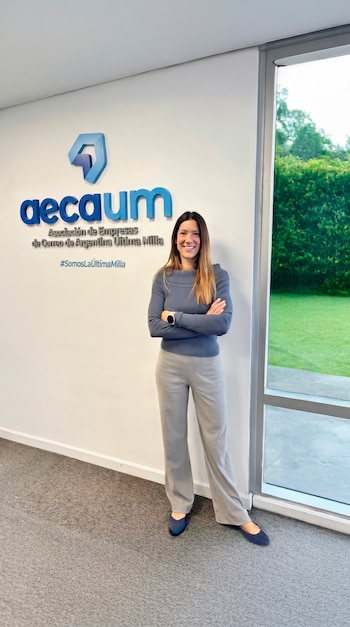 Una mujer de cabello castaño con blusa azul y pantalón gris claro sonríe con los brazos cruzados frente a un logo de AECAUM y una ventana a un jardín verde