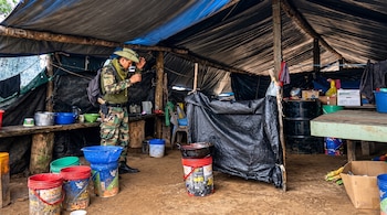 Un hombre con uniforme en un cobertizo de lonas con baldes de plástico, ollas de cocina, barriles y suelo de tierra.