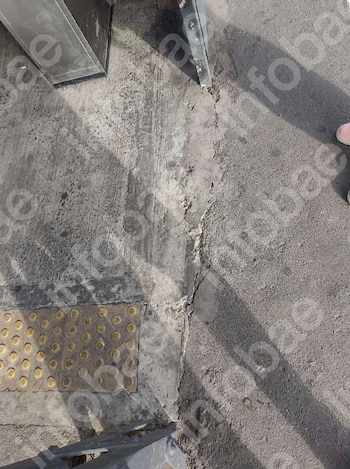 Suelo de concreto gris con grietas y parches irregulares en una estación de tren. Incluye una franja de pavimento táctil amarillo y partes de una estructura metálica