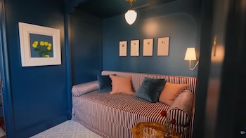 Habitación con paredes azul oscuro, un daybed a rayas con cojines azules y rosas. Cuadros en la pared, una lámpara de pared. Mesita de noche con una rosa
