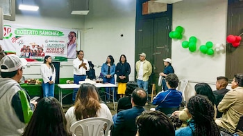 Un hombre de camisa blanca y gafas habla ante una audiencia sentada, con un banner político y personas de pie detrás. La pared tiene globos verdes y rojos