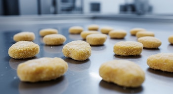 Nuggets congelados esparcidos sobre una superficie brillante de acero inoxidable en un entorno industrial con iluminación fría y fondo borroso.