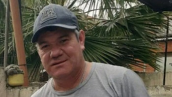 Primer plano de un hombre de mediana edad con gorra azul, camiseta gris y cadena, posando frente a follaje verde y una pared con planta en maceta