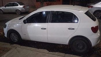 Un coche hatchback blanco de cuatro puertas está estacionado en una calle oscura, visto desde el lado. Otro coche plateado se ve al fondo