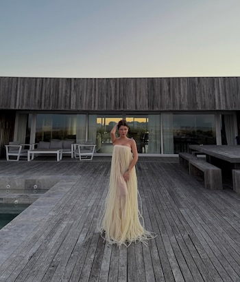 Una mujer de pie con un vestido largo y claro, un deck de madera, una piscina, una casa moderna con ventanales y muebles de exterior, cielo claro