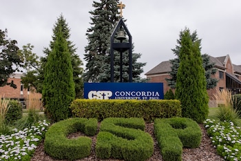 Un cartel azul con el texto Concordia University, una campana con cruz, arbustos podados con las letras CSP, abetos y flores