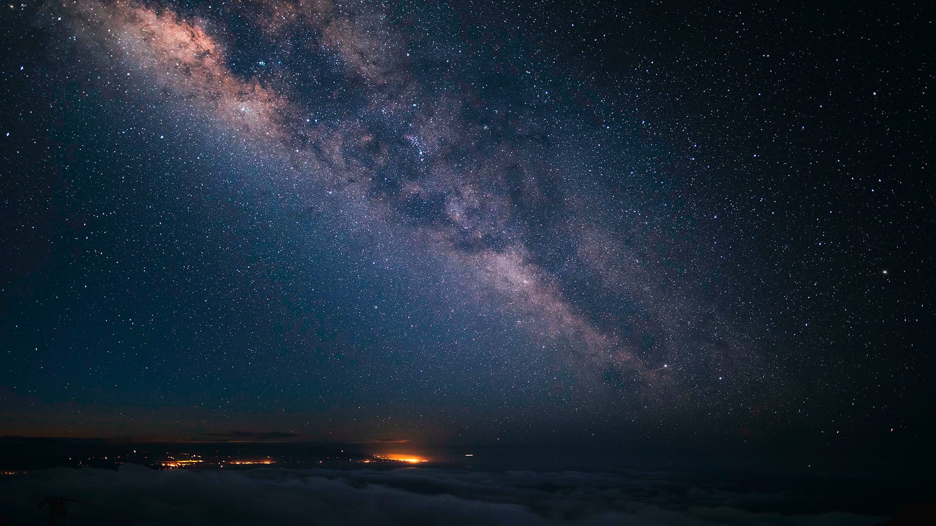La Vía Láctea cruza el cielo nocturno sobre un mar de nubes y luces distantes en la cumbre del volcán Mauna Kea, en Hawái. (Pexels)