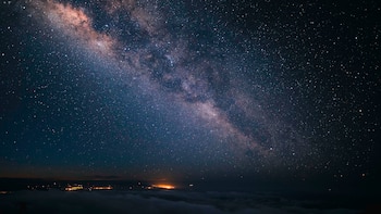 Cielo nocturno estrellado atravesado en diagonal por la Vía Láctea, sobre una capa de nubes oscuras y luces en el horizonte