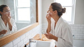 Una mujer con bata de baño blanca aplica crema facial en su rostro frente a un espejo, con un lavabo y una ventana al fondo.
