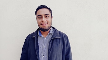 Hombre joven con cabello oscuro y barba, camisa de rayas, chaqueta oscura, sonriendo. Fondo de pared blanca texturizada.