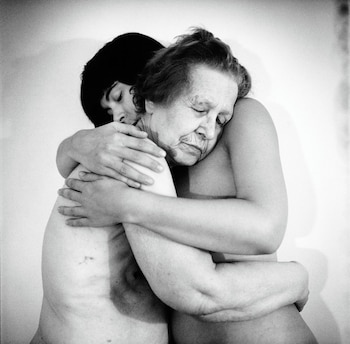 Dos mujeres desnudas de distintas edades se abrazan con los ojos cerrados en una fotografía en blanco y negro frente a un fondo claro