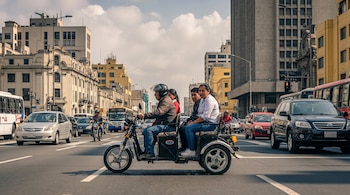 Trimoto eléctrica negra con conductor y dos pasajeros circula en calle urbana. Otros vehículos y edificios componen el entorno con cielo parcialmente nublado.
