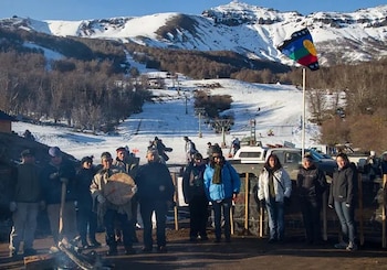 Grupo de personas Mapuche alrededor de una fogata, con un tambor, frente a pistas de esquí nevadas y un remonte en el Cerro Chapelco, bajo un cielo azul. Una bandera Mapuche flamea a la derecha