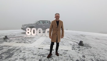 Hombre con abrigo y gafas de pie sobre una superficie nevada, un vehículo utilitario gris y números grandes blancos '800.000' en el fondo