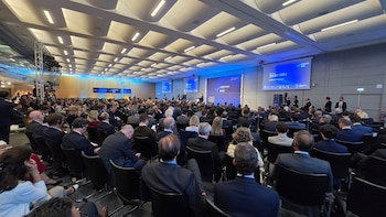 Un salón de conferencias lleno de asistentes sentados frente a un escenario con pantallas azules que muestran el nombre de Javier Milei