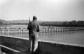 Fotografía en blanco y negro de un hombre de espalda con uniforme militar y fusil, junto a un muro bajo frente a un río con un puente de madera y montañas
