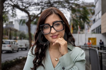 Mujer con cabello oscuro, gafas de montura negra, blusa verde claro y collar dorado, con una mano en la barbilla, al aire libre con fondo urbano borroso