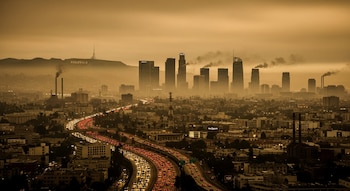 Vista aérea de Los Ángeles con un cielo brumoso y anaranjado, rascacielos, chimeneas industriales emitiendo humo y una autopista congestionada de vehículos.