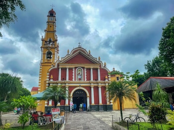 Iglesia de fachada roja y amarilla con campanario y reloj bajo cielo nublado. Palmeras, árboles, personas y una bicicleta visibles en el exterior