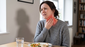 Una mujer sentada a la mesa se toca el cuello, que está resaltado en rojo, frente a un plato con vegetales y un vaso con agua.