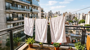 Toallas blancas sujetadas con broches de madera en una soga extendida en el balcón de un edificio, con macetas y la ciudad al fondo.