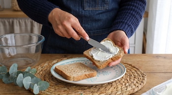 Manos de una persona untando queso crema en una rebanada de pan integral con un cuchillo. Una rebanada de pan en un plato, un cuenco de cristal, hojas de eucalipto y un tapete