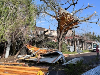 Calle con árboles caídos y ramas. Escombros de techos y maderas en el suelo. Una estructura de chapa y madera en un árbol. Personas observando los daños. Edificios residenciales y cielo azul