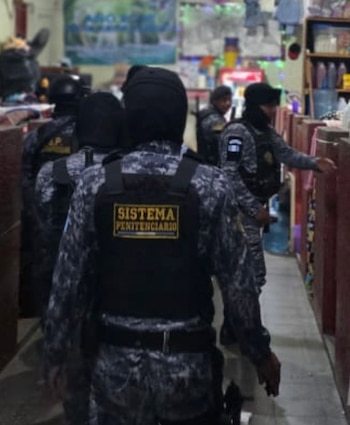 Varias personas uniformadas con camuflaje, chalecos negros con "Sistema Penitenciario", gorras oscuras, en pasillo interior con estantes