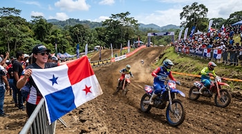 Una mujer sostiene la bandera de Panamá junto a una pista de tierra donde varios pilotos compiten en motocross ante el público.