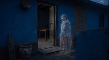 Figura translúcida de una mujer anciana de pie ante la puerta abierta de una casa azul de adobe, con una mesa al fondo y pétalos en el suelo.