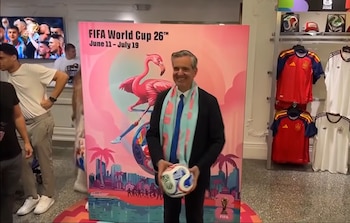 Un hombre sonriente en traje y bufanda azul sostiene un balón de fútbol oficial frente a un gran cartel rosa de la Copa Mundial 2026 en el interior de una tienda con camisetas y balones
