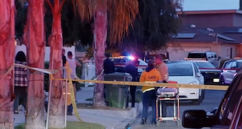 Escena nocturna de un crimen con cinta policial amarilla, personal de emergencia LAFD y policías, y coches con luces intermitentes en una calle residencial. Se ven palmeras y casas