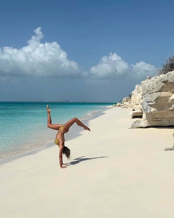 Martina Díaz Radonic, parada de manos sobre la arena húmeda de la orilla, con las piernas abiertas en el aire y un bikini amarillo mostaza. Las formaciones rocosas y el agua turquesa se extienden detrás de ella