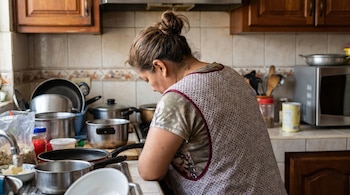 Vista trasera de una mujer latina de mediana edad con el cabello recogido y delantal, apoyada en el mostrador de una cocina con utensilios y ollas.