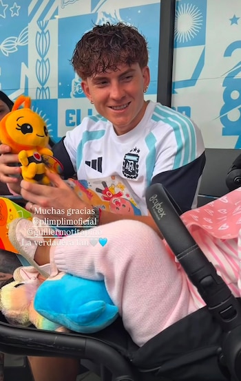 Hombre vestido con camiseta de fútbol de Argentina sostiene un muñeco de abeja. Piernas de bebé con pantalón rosa en un cochecito. Varios juguetes de peluche