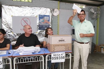José Williams sonríe mientras sostiene una boleta de votación y un bolígrafo sobre una urna de ONPE, con personal electoral sentado detrás