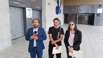 Tres personas de pie frente a la entrada de un edificio público mientras el hombre del centro sostiene un documento impreso